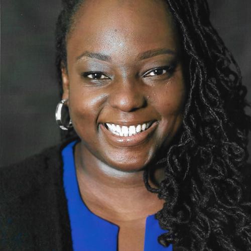 A portrait of Denisha Jones smiling with silver earrings. Curly hair cascades down the right side. She is wearing a blue dress with a black jacket over top. 