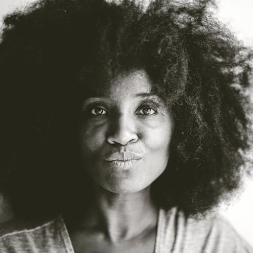 black and white headshot of a smiling black woman with a large afro and a grey v neck jersey top who happens to be fashion designer, costume designer, writer, filmmaker, visual artist and college professor Maria Fenton