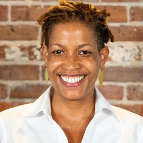 A headshot of a middle-aged Black woman with short locs. She is smiling warmly. She is wearing a white dress shirt and the background features a textured brick wall.