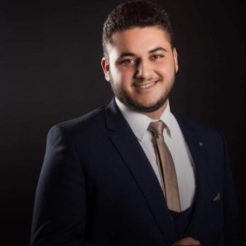 A young man with short dark hair and a trimmed beard, wearing a dark navy blue suit, light dress shirt, and beige tie. He is smiling and standing in front of a plain black background in a professional studio portrait.