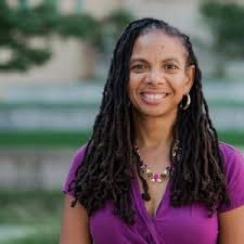 A Black woman with locs who is smiling and wearing a purple blouse.