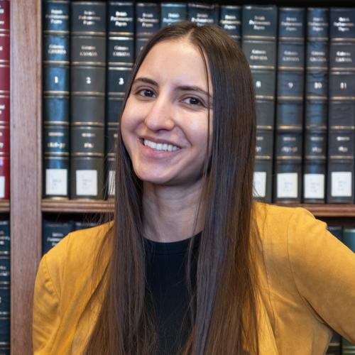 picture of a white woman from the chest up. She has long brown hair, brown eyes and is smiling. She is standing in front of a book case.