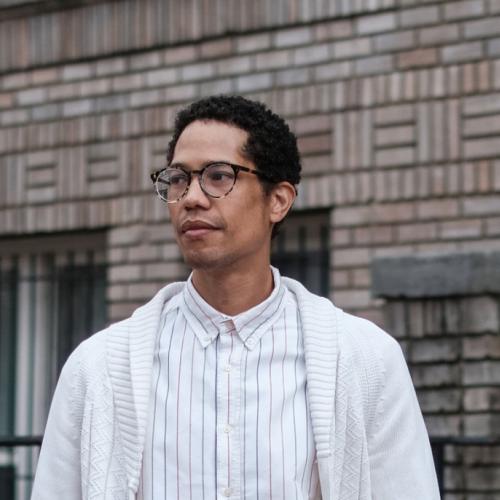 Person wearing light colored shirt and sweater standing outside. Visible from chest up. They are also wearing glasses.