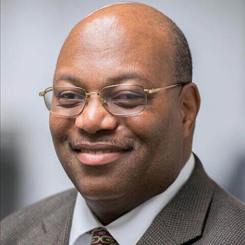 John Gordon, smiling and wearing glasses with a white button down shirt, tie and suit jacket.