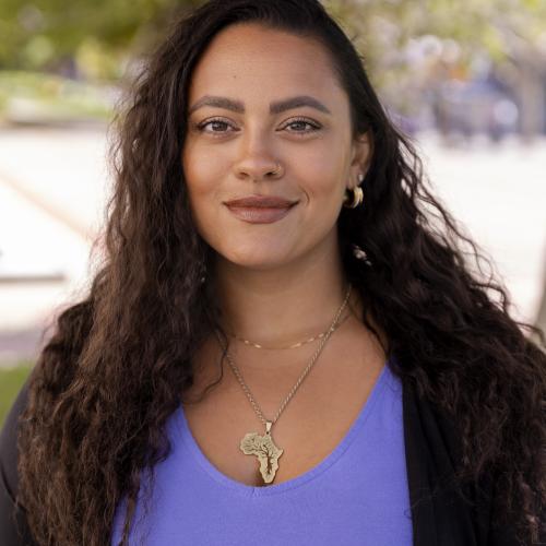 The image depicts Kelekay standing on a campus yard, wearing a purple top and black blazer.