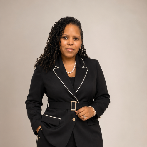Dominique Douglas standing against a beige background, wearing a black pantsuit with white piping